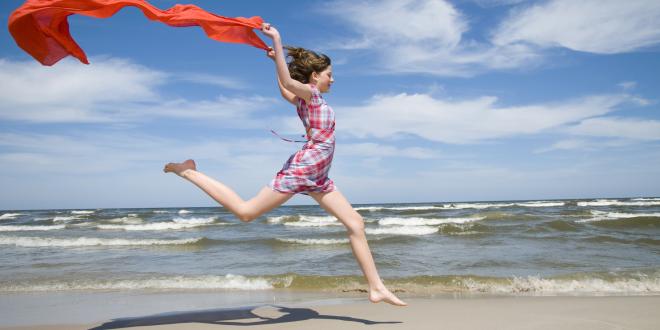 A teenage girl runs along a beach trailing a red cloth behind her.